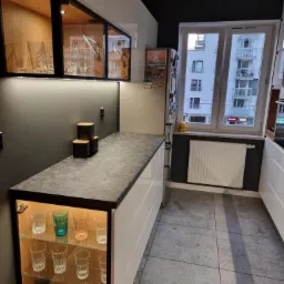 Nowoczesna kuchnia z białymi szafkami, szarym blatem i podświetlanymi witrynami z kolekcją szklanek, widok na budynek mieszkalny przez okno.