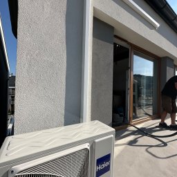 ClimJack Jacek Nosek - Zewnętrzna jednostka klimatyzacji Haier na balkonie z widoczną elewacją budynku i pracownikiem w tle podczas instalacji.