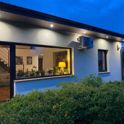 ClimJack Jacek Nosek - Biały budynek jednorodzinny z zamontowaną jednostką klimatyzacji zewnętrznej marki Haier, widoczne oświetlenie zewnętrzne i okno z widokiem na schody wewnętrzne i rośliny doniczkowe, krzewy przed...