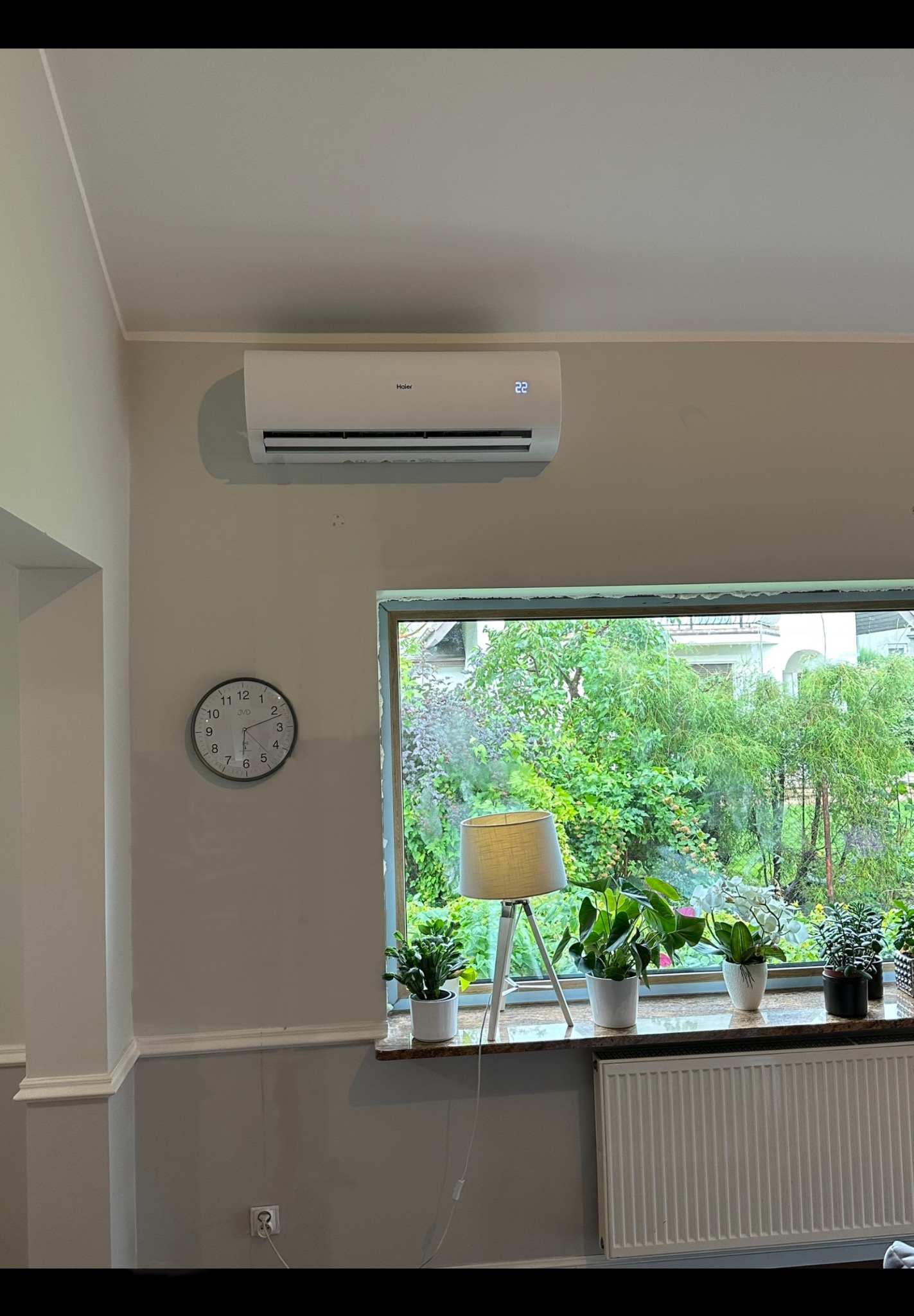 Biały klimatyzator Haier zamontowany na ścianie w jasnym pokoju, obok okna z widokiem na zielony ogród, zegar na ścianie.