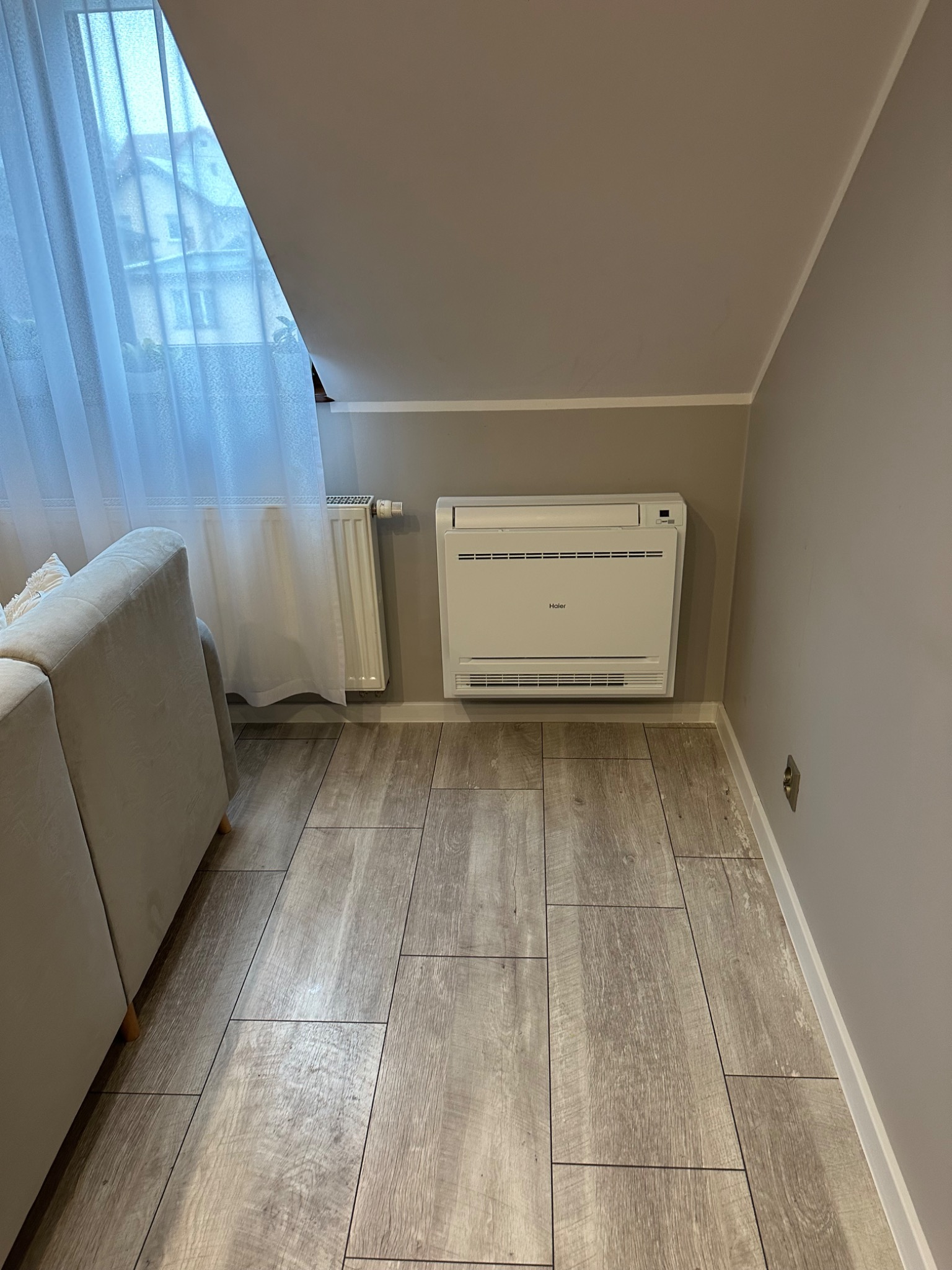 Biały klimatyzator Haier zamontowany na ścianie w pokoju na poddaszu, obok grzejnika i okna zasłoniętego białą firanką, na podłodze panele imitujące drewno.