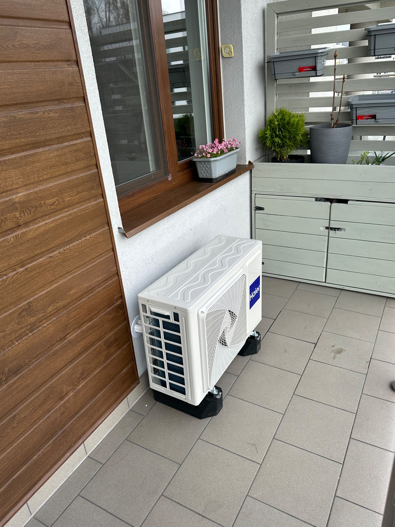 Biała jednostka zewnętrzna klimatyzacji Haier umieszczona na czarnych podkładkach antywibracyjnych, stoi na szarej, kafelkowej podłodze balkonu, obok brązowej ściany z sidingu i skrzynki z kwiatami.