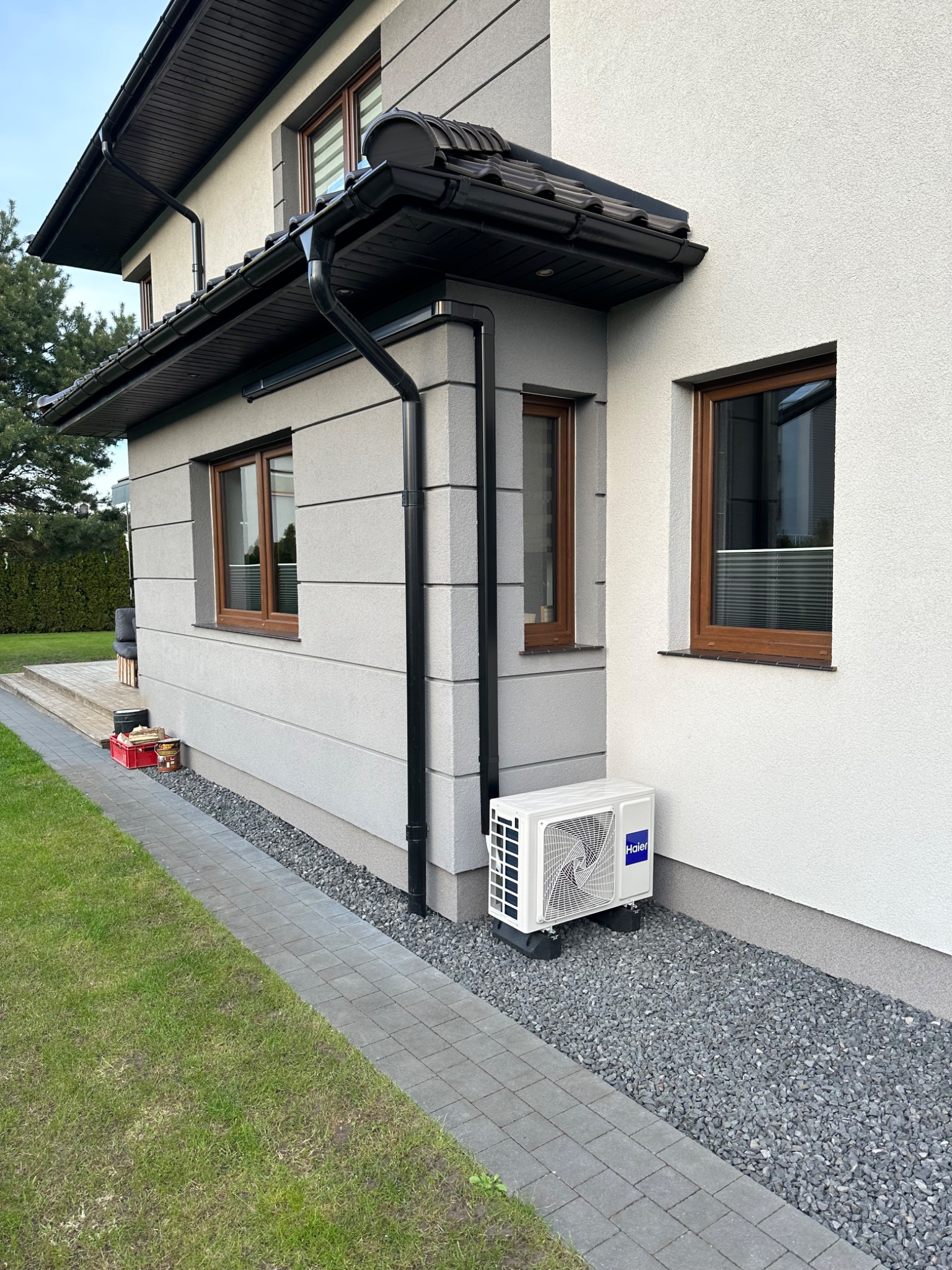 Biała jednostka zewnętrzna klimatyzacji Haier zamontowana na szarym żwirze przy ścianie domu w Redzie. Widoczna elewacja z oknami w brązowych ramach i ciemnymi rynnami.