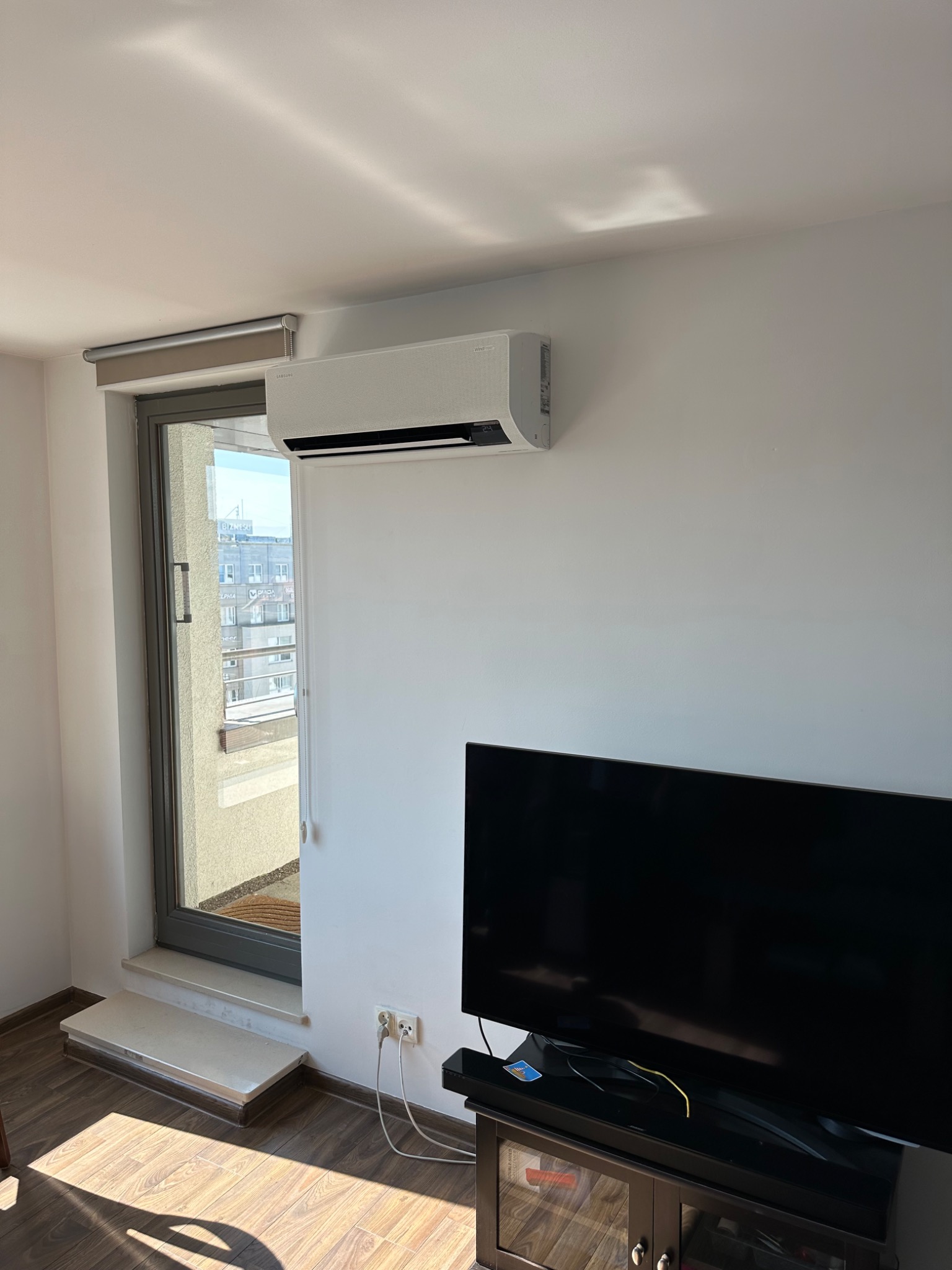 Biały klimatyzator ścienny zamontowany nad telewizorem w jasnym pokoju z balkonem, widoczne kable zasilające.