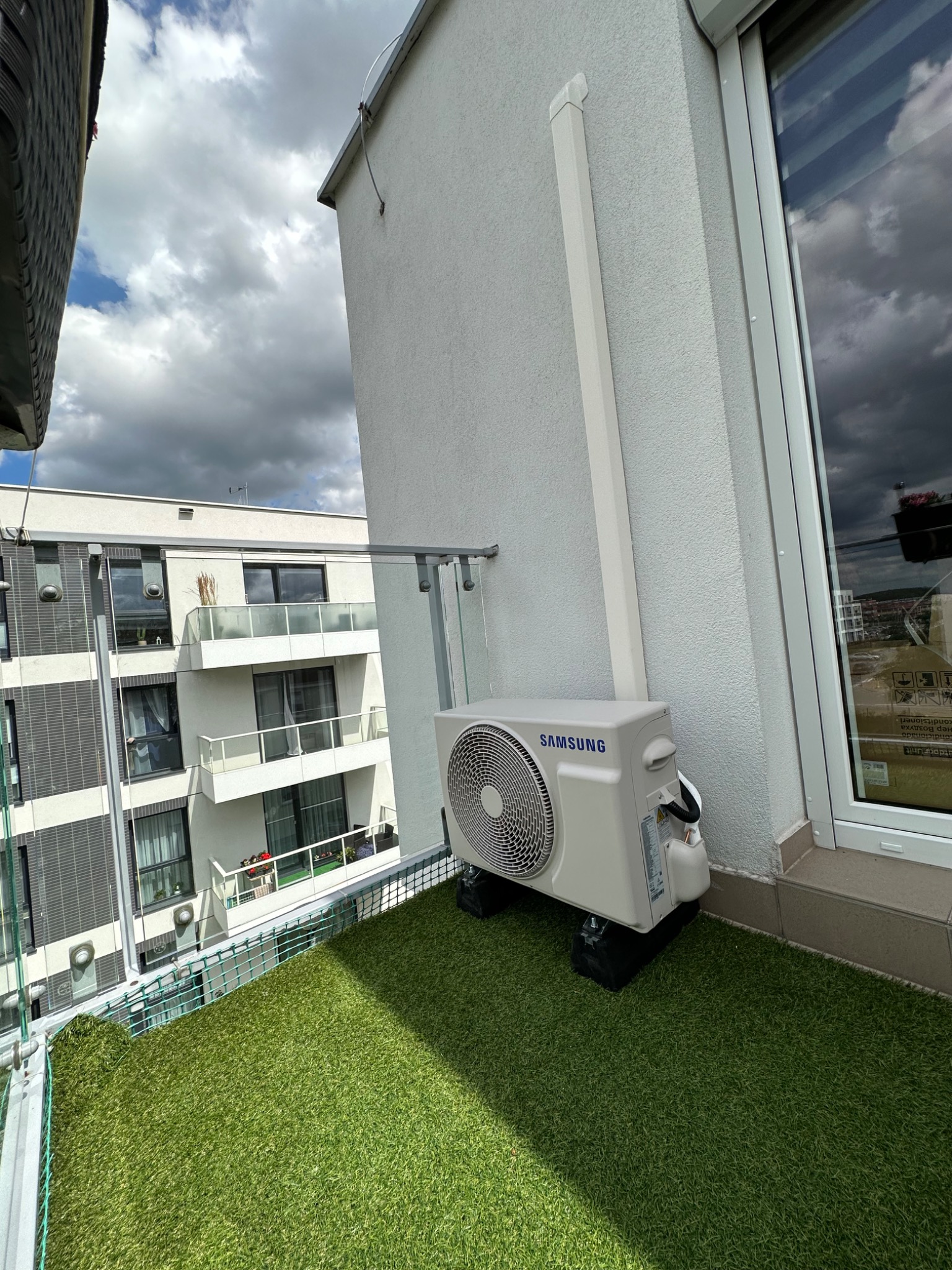 Jednostka zewnętrzna klimatyzatora Samsung zamontowana na balkonie z sztuczną trawą, widoczny budynek mieszkalny w tle i rura odprowadzająca skropliny.