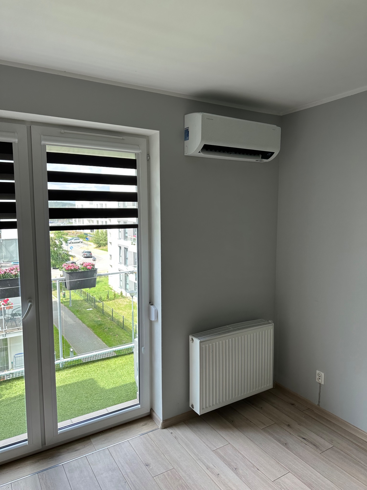 Zamontowany klimatyzator Samsung na szarej ścianie w pokoju z grzejnikiem i widokiem na balkon z kwiatami.