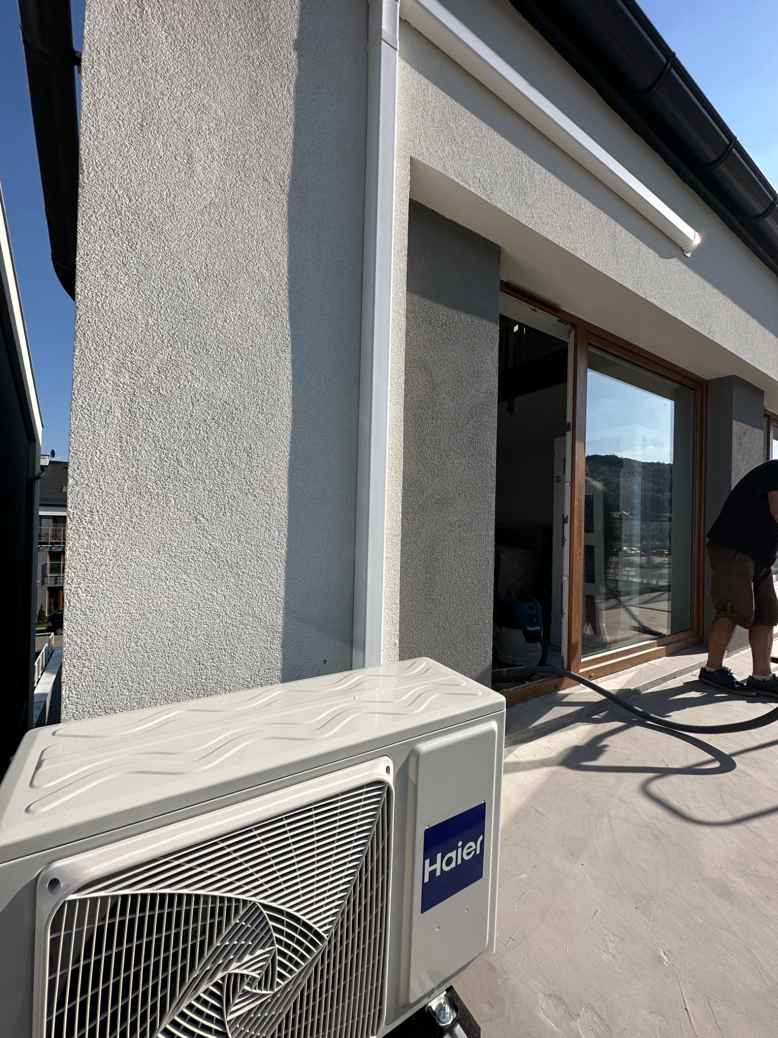 Zewnętrzna jednostka klimatyzacji Haier na balkonie z widoczną elewacją budynku i pracownikiem w tle podczas instalacji.