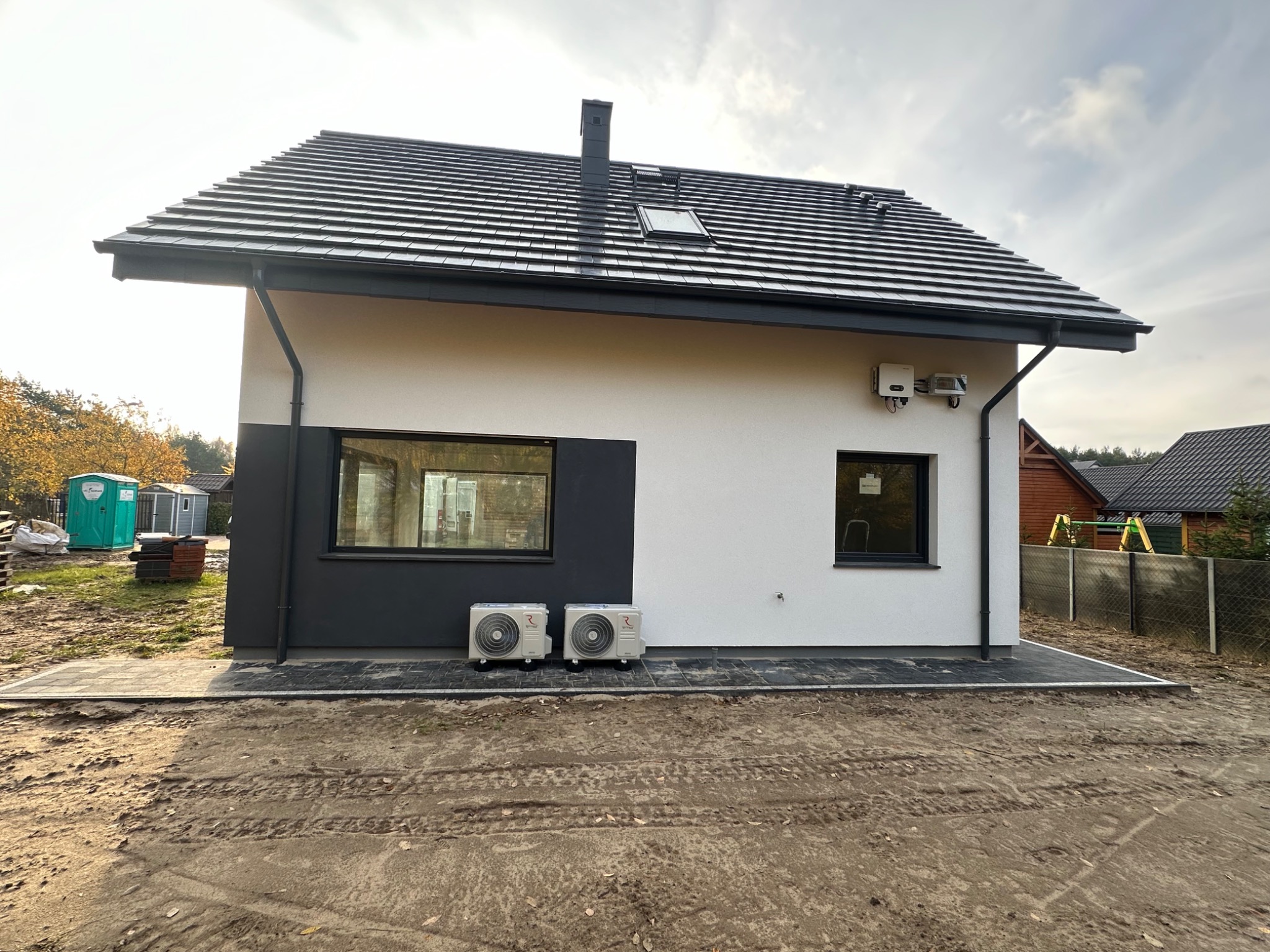 Dwa zewnętrzne agregaty klimatyzacji zamontowane na kostce brukowej przy ścianie nowoczesnego domu z dwukolorową elewacją, widoczne także skrzynki elektryczne na ścianie.