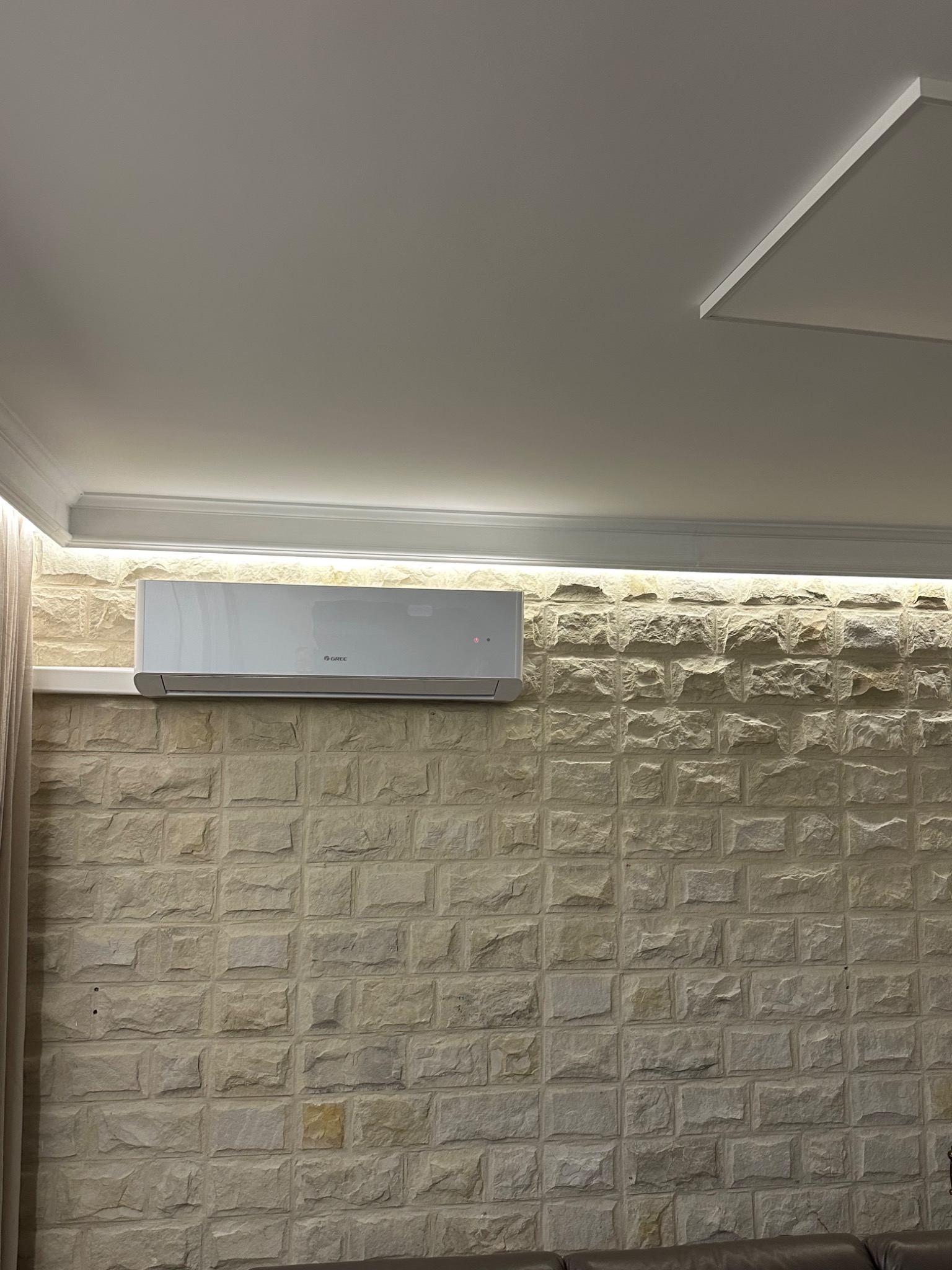 Biały klimatyzator marki Gree zamontowany na ścianie wyłożonej dekoracyjną cegłą, z oświetleniem LED pod sufitem.