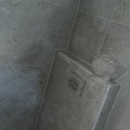 Mateusz Pfeiffer - Szare płytki w łazience z zabudowaną spłuczką WC, częściowo zabezpieczoną folią. Widoczne wystające rury instalacji wodnej. Minimalistyczny design.