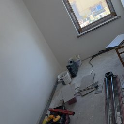 Mateusz Pfeiffer - Wnętrze w trakcie remontu: okno dachowe, białe ściany, narzędzia glazurnicze (przecinarka, poziomica), płytki i wiadro z zaprawą na podłodze.