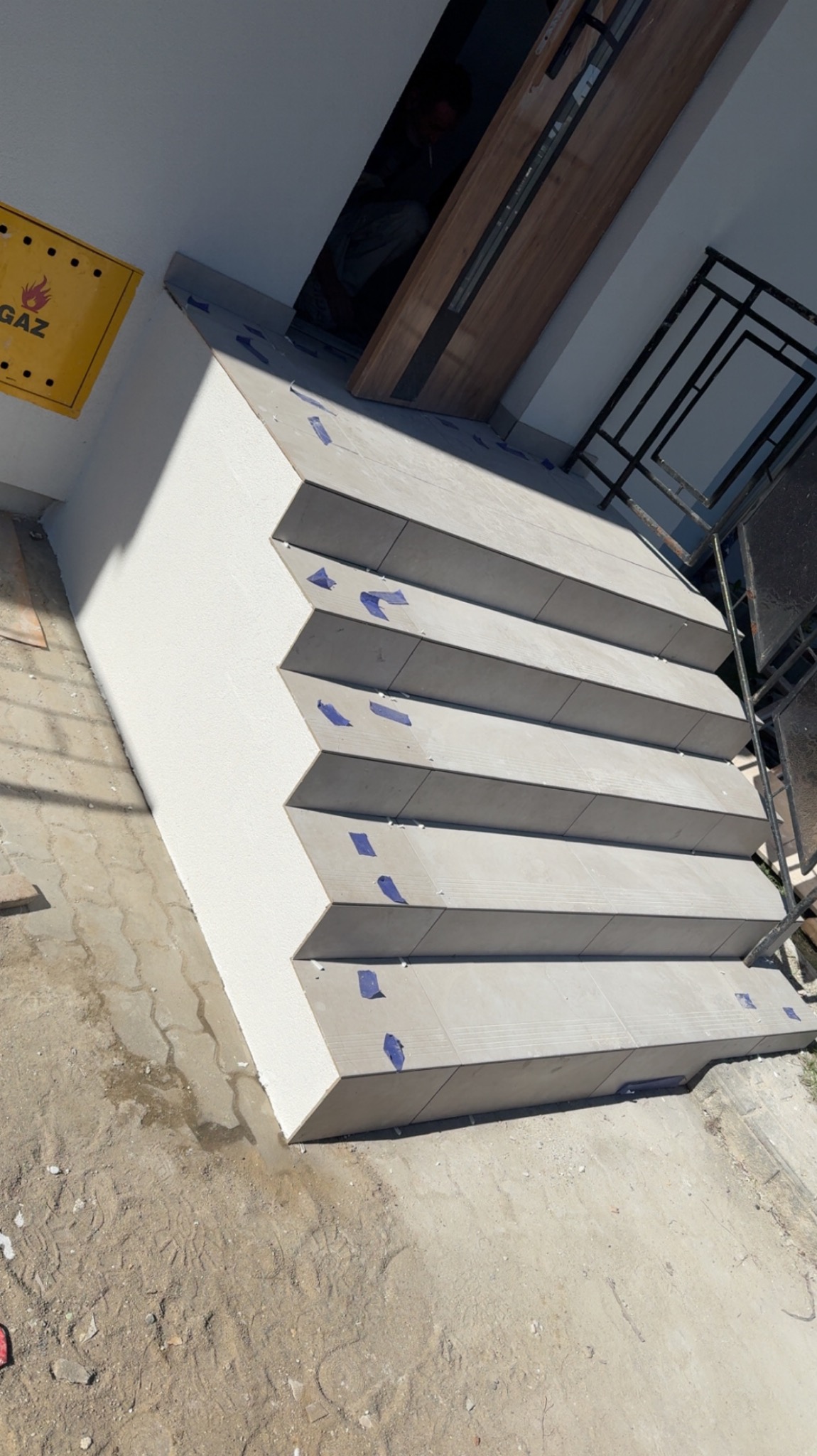 Schody wyłożone szarymi płytkami z niebieską taśmą zabezpieczającą, jasna ściana, fragment balustrady i żółta skrzynka gazowa w tle. Kątowe ujęcie.