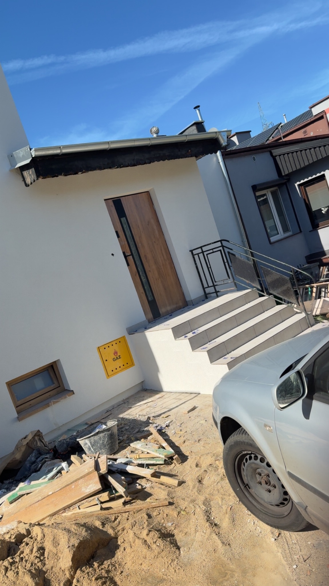 Nowo otynkowany dom jednorodzinny z drewnianymi drzwiami wejściowymi, schodami z metalową barierką i żółtą skrzynką gazową. Na ziemi gruz i piasek.