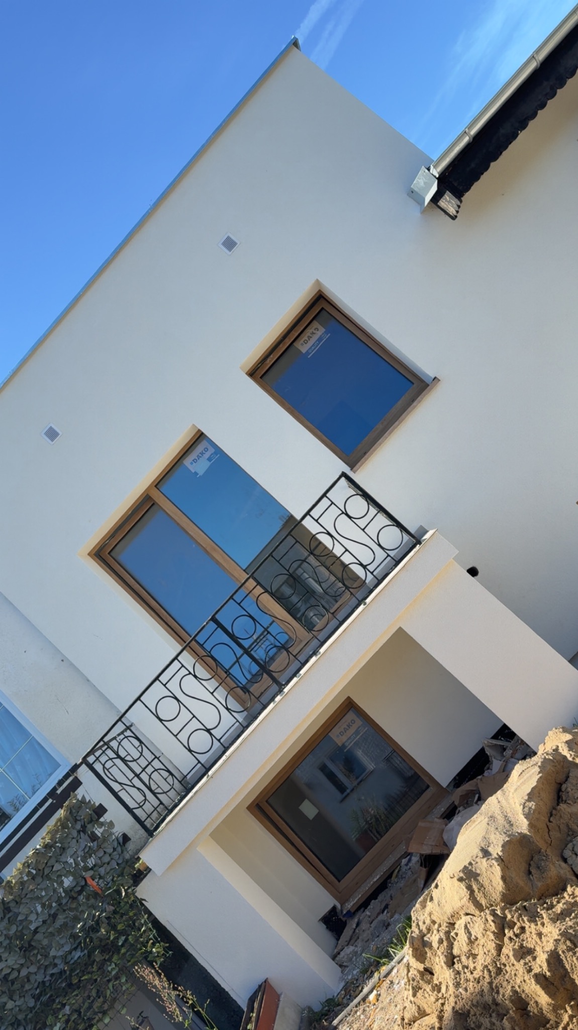 Biała elewacja domu z oknami w drewnianych ramach, balkonem z ozdobną balustradą i fragmentem niezagospodarowanego terenu. Widoczne naklejki na szybach okien.
