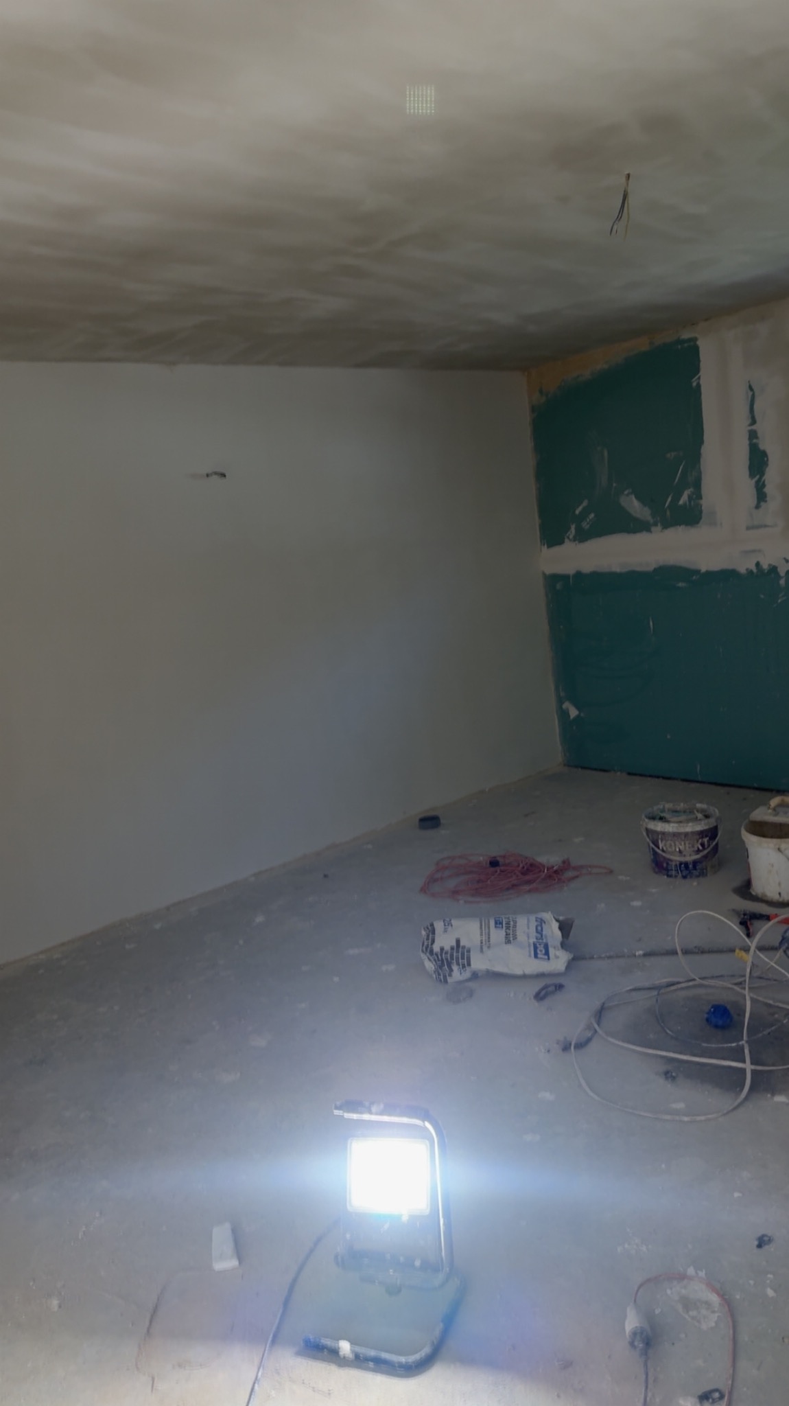 Pokój w trakcie prac wykończeniowych: szpachlowane ściany i sufit, płyty karton-gips na ścianie, narzędzia i lampa budowlana na podłodze. Widok na wnętrze w trakcie remontu.