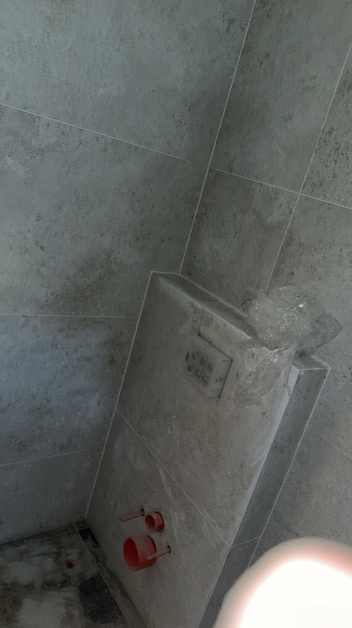 Szare płytki w łazience z zabudowaną spłuczką WC, częściowo zabezpieczoną folią. Widoczne wystające rury instalacji wodnej. Minimalistyczny design.