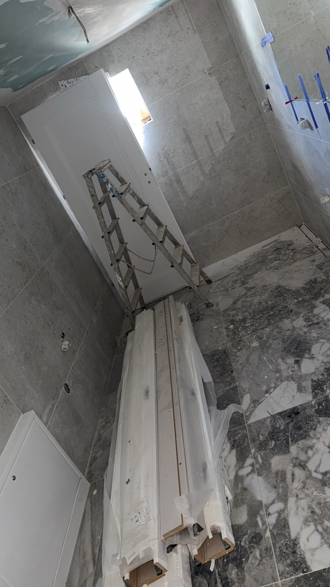 Montaż sufitu podwieszanego w łazience z marmurową podłogą. Widoczna drabina, płyta gipsowa i profile w folii. Ściany wykończone szarą płytką.