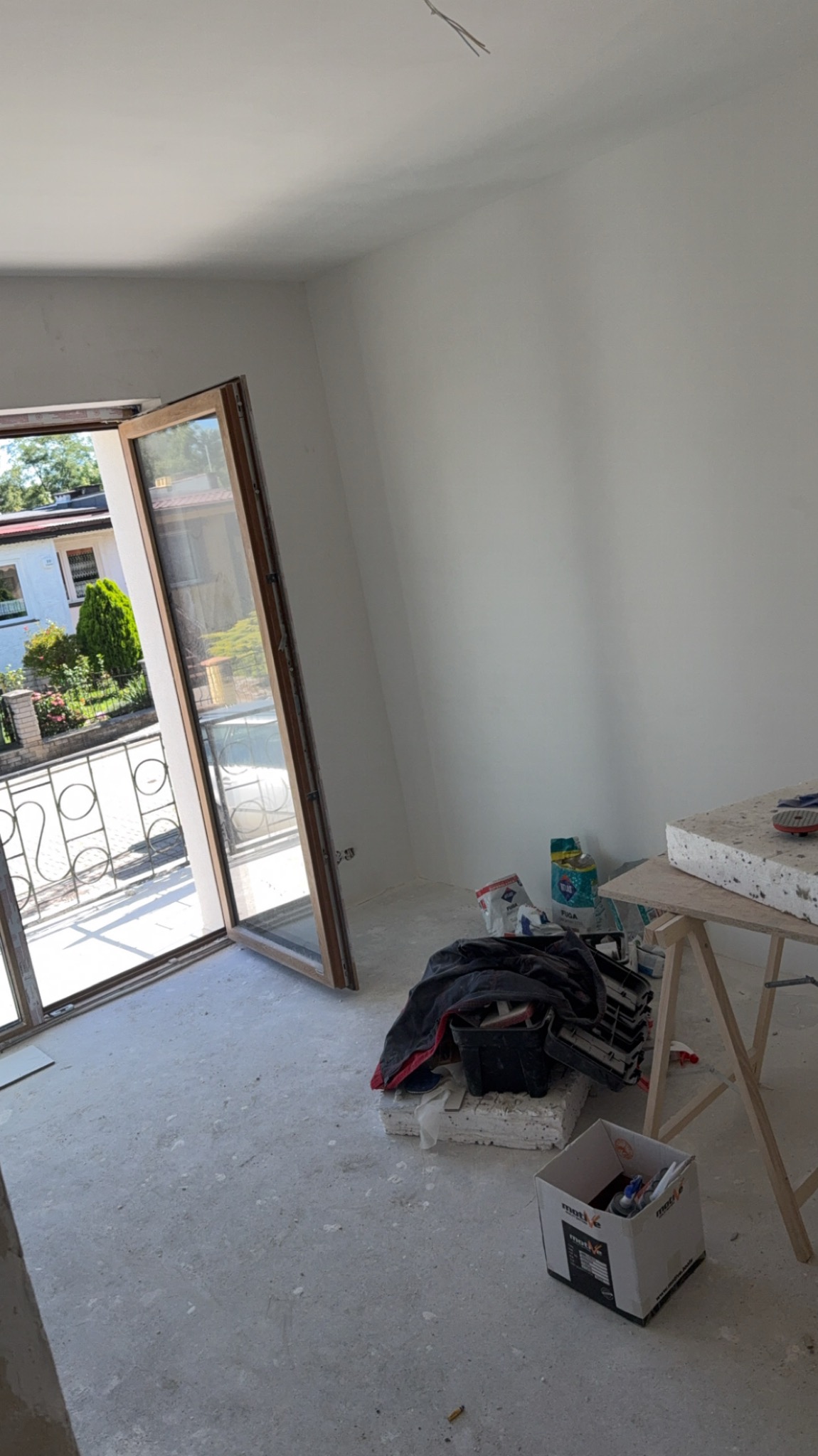 Wnętrze pokoju w trakcie prac wykończeniowych: świeżo pomalowane ściany, otwarte okno balkonowe i narzędzia malarskie na podłodze, widok na zieleń za oknem.