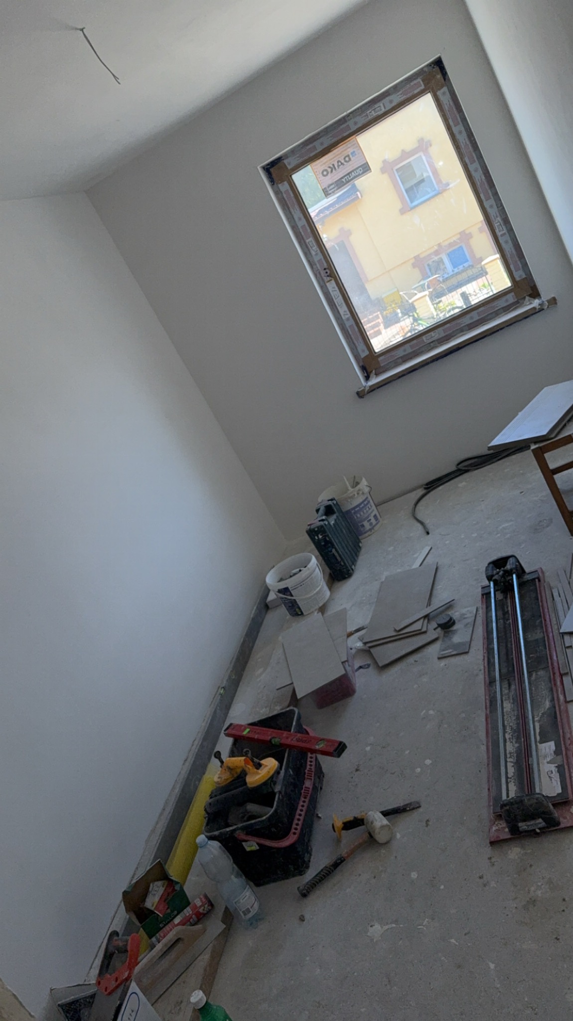 Wnętrze w trakcie remontu: okno dachowe, białe ściany, narzędzia glazurnicze (przecinarka, poziomica), płytki i wiadro z zaprawą na podłodze.