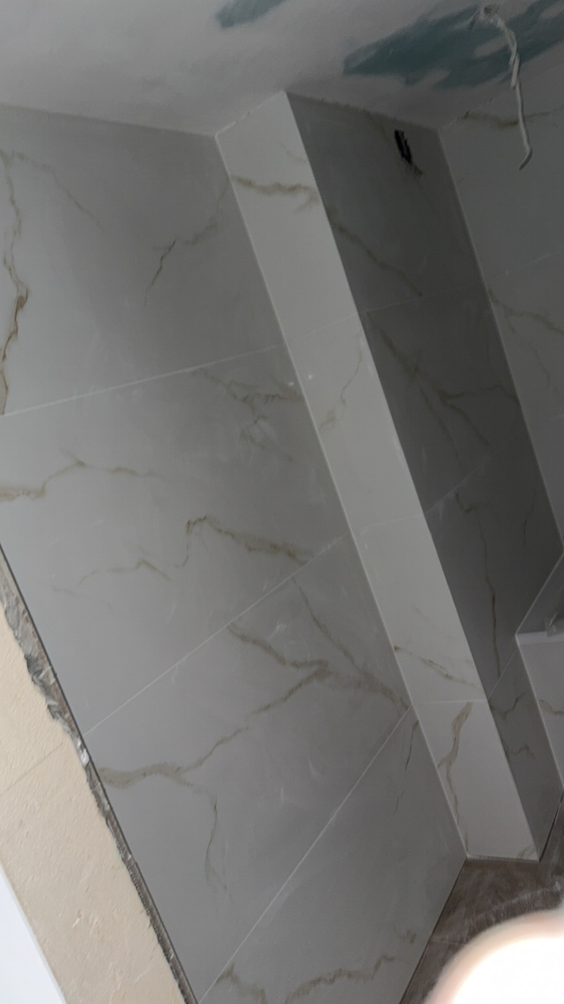 Ściana wyłożona białymi płytkami imitującymi marmur, z widocznym fragmentem sufitu i narożnika, w trakcie prac wykończeniowych. Widoczny kabel elektryczny.