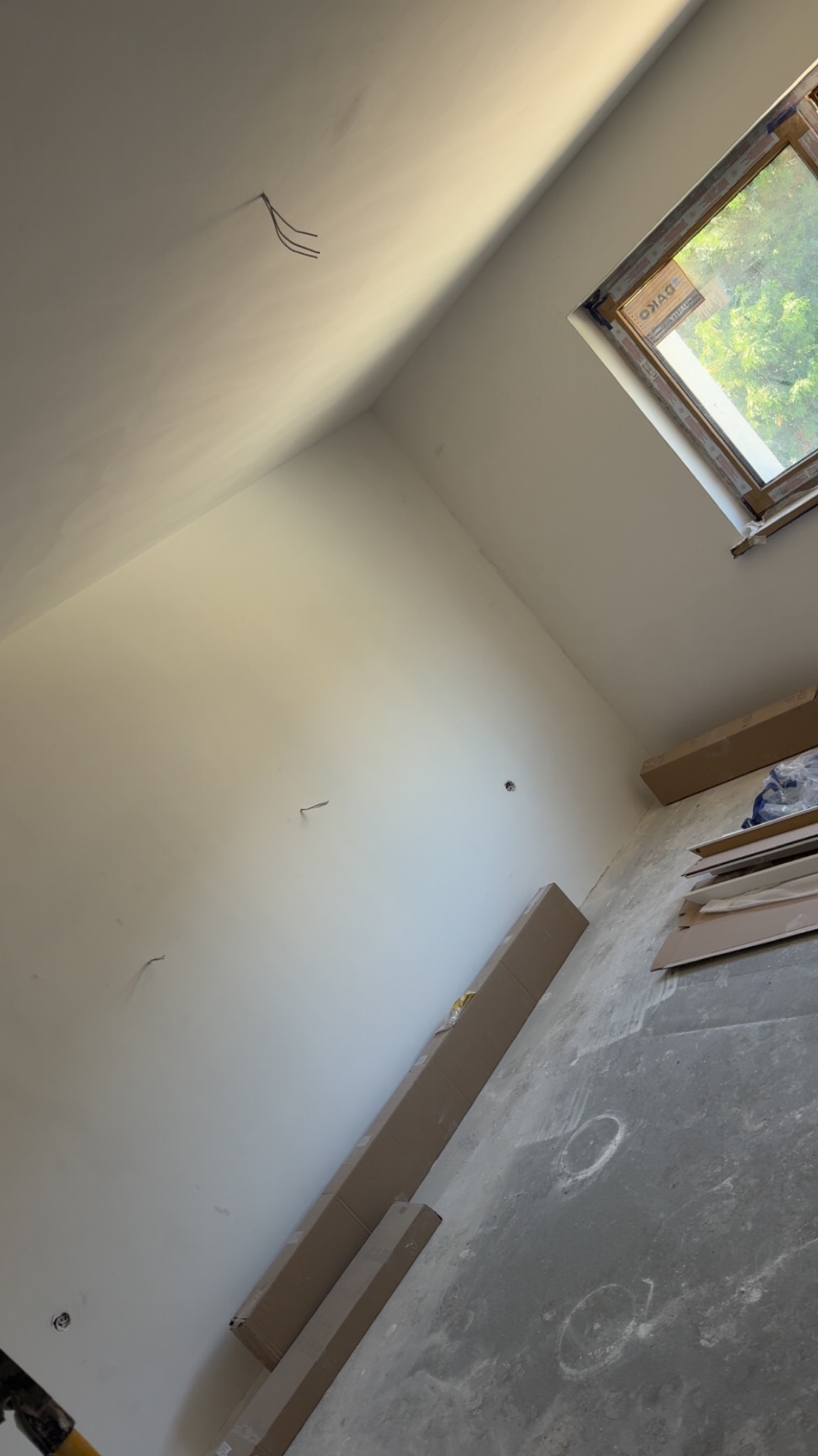 Wnętrze poddasza po szpachlowaniu ścian i sufitu, widoczne kable, okno dachowe i materiały budowlane na podłodze. Przygotowanie do dalszych prac wykończeniowych.