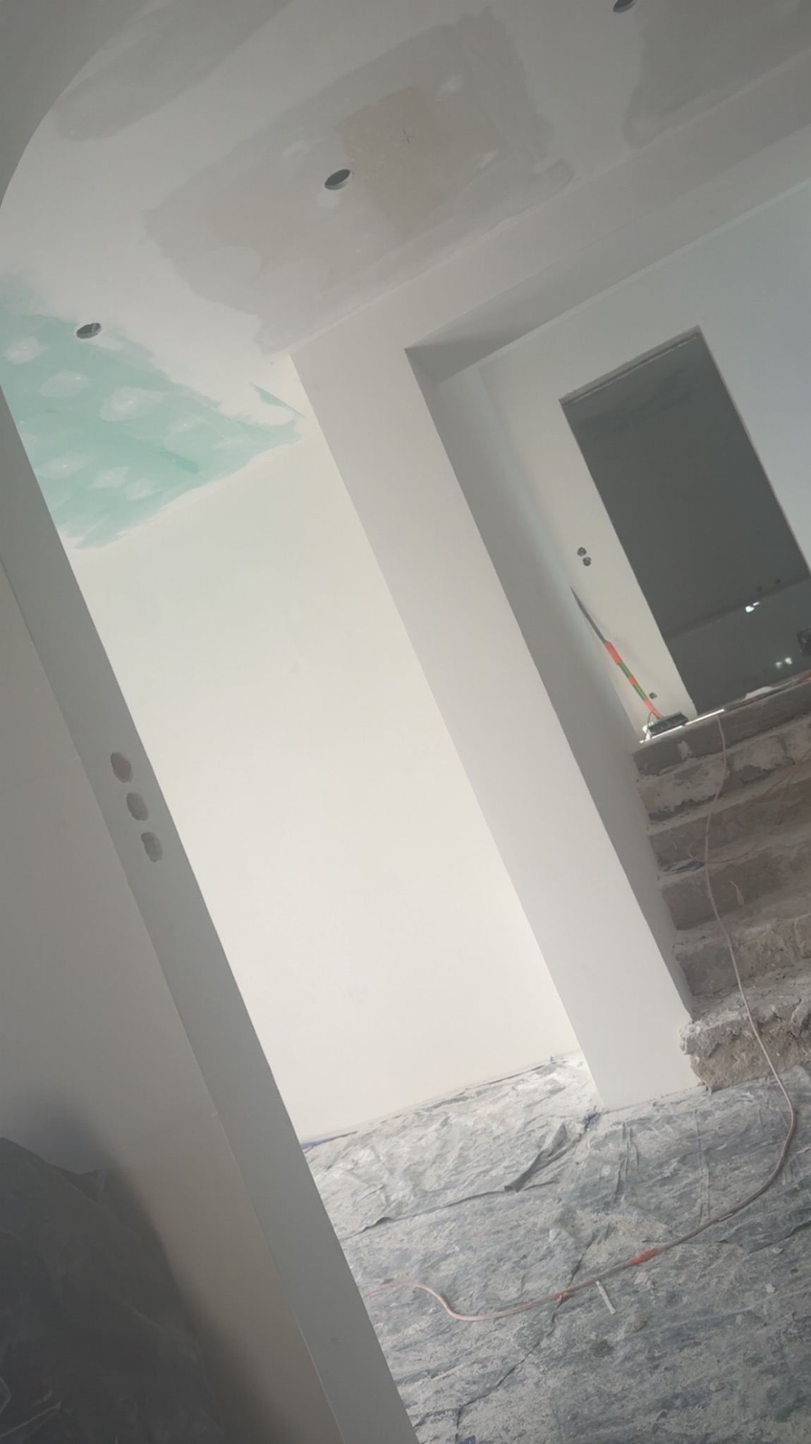 Wnętrze w trakcie malowania: białe ściany, sufit z zielonymi śladami farby, widoczne otwory na oświetlenie, zabezpieczona podłoga, fragment schodów w tle.