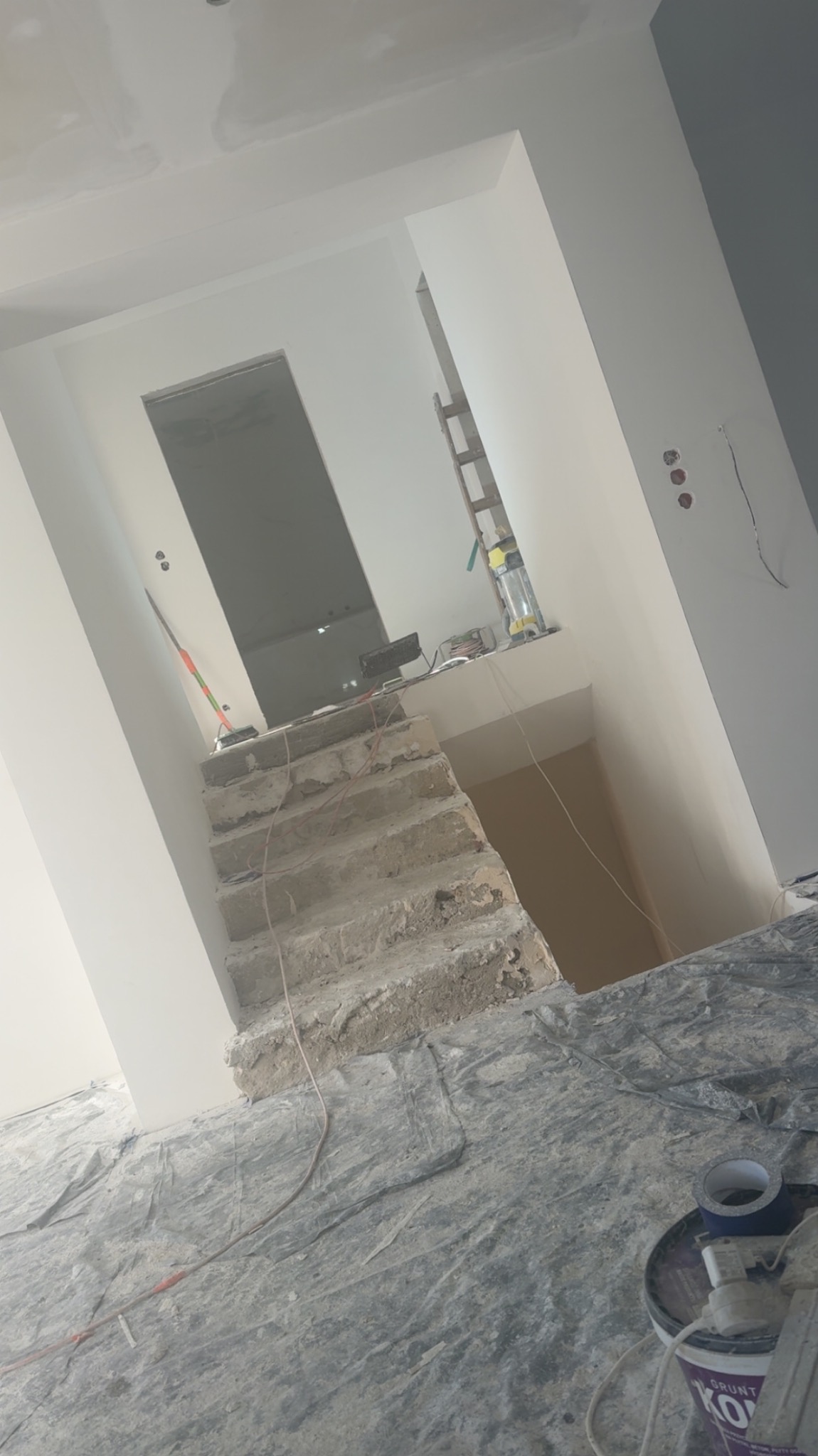 Remont wnętrza: surowe schody, świeżo pomalowane ściany na biało, zabezpieczona folią podłoga i wiadro z gruntem. Widoczne niedoskonałości i etap prac.