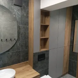 Nowoczesna łazienka z szarymi płytkami imitującymi marmur, drewnianym blatem pod umywalką nablatową, okrągłym lustrem z czarnymi haczykami i szarą zabudową meblową z drewnianymi akcentami...
