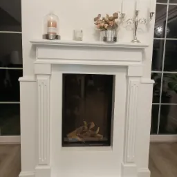 Biały, klasyczny portal kominkowy z dekoracjami: świecą pod kloszem, świecznikiem i bukietem, na tle okna z podziałami.