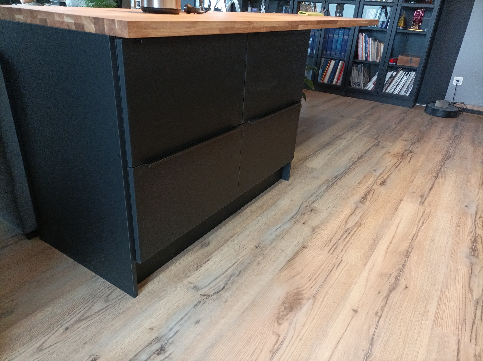 Wyspa kuchenna z czarnymi szafkami i drewnianym blatem, widok z boku, na podłodze panele, w tle regał z książkami i robot sprzątający.