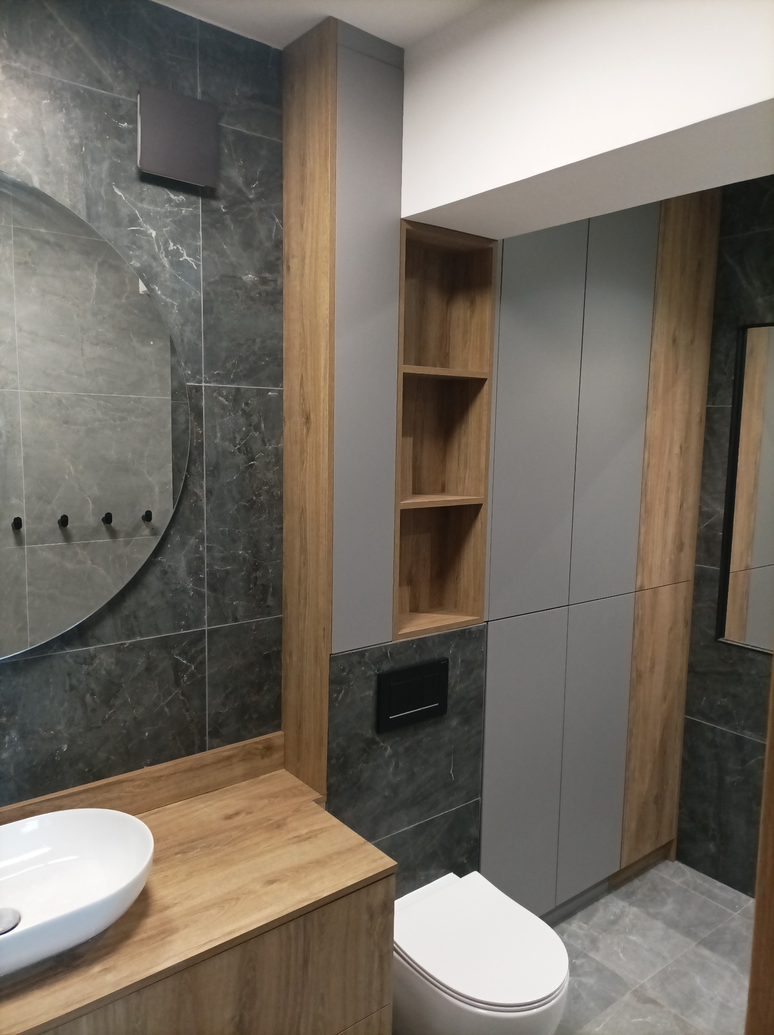 Nowoczesna łazienka z szarymi płytkami imitującymi marmur, drewnianym blatem pod umywalką nablatową, okrągłym lustrem z czarnymi haczykami i szarą zabudową meblową z drewnianymi akcentami...