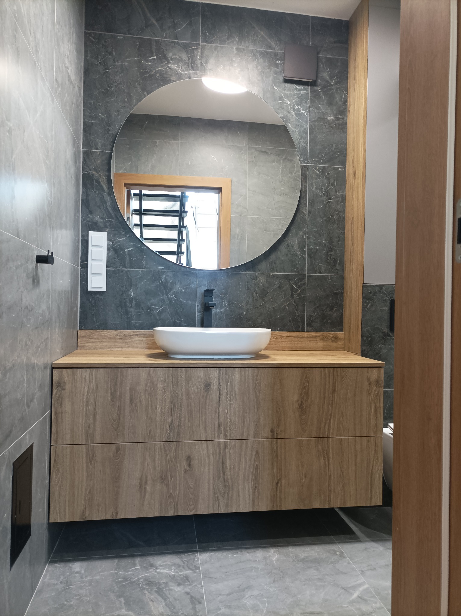 Nowoczesna łazienka z szarą marmurową okładziną, okrągłym lustrem z podświetleniem, białą umywalką nablatową na szafce z drewnopodobnym fornirem i czarną armaturą.