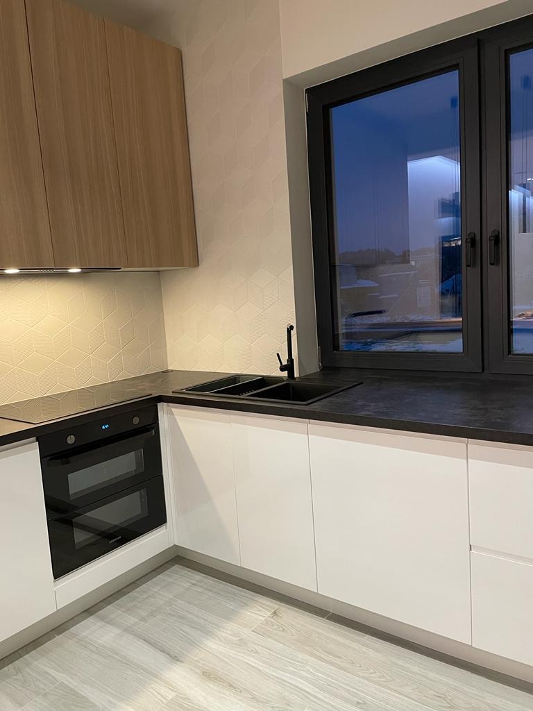 Nowoczesna kuchnia z białymi frontami szafek, drewnianymi szafkami wiszącymi, czarnym blatem i zlewozmywakiem, oraz dużym oknem z widokiem na basen.