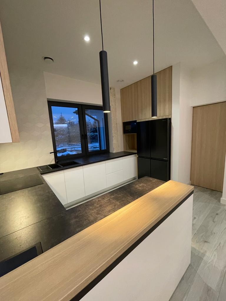 Nowoczesna kuchnia z wyspą, białe fronty szafek, czarny blat i lampy tuby, widok na zimowy krajobraz za oknem.