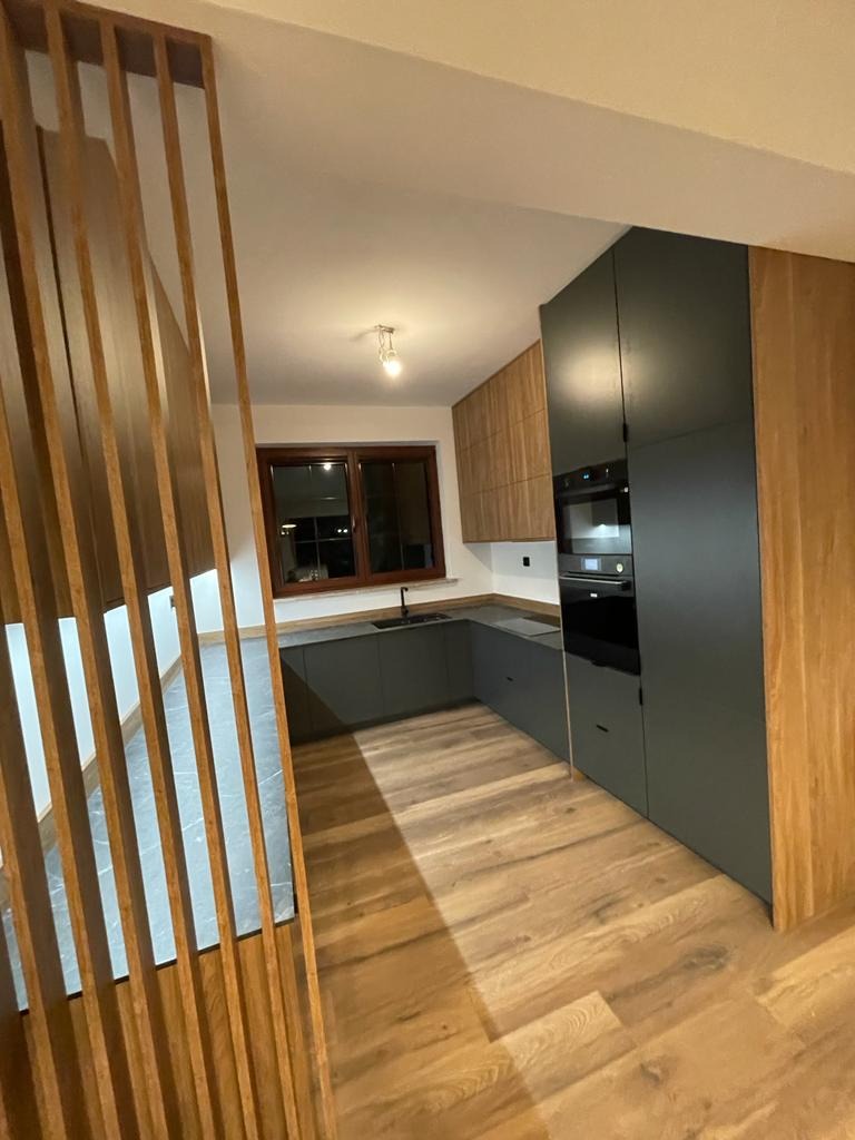 Nowoczesna kuchnia z szarymi szafkami dolnymi i drewnianymi górnymi, oddzielona drewnianą ażurową ścianką działową, z widocznym oknem i drewnianą podłogą.