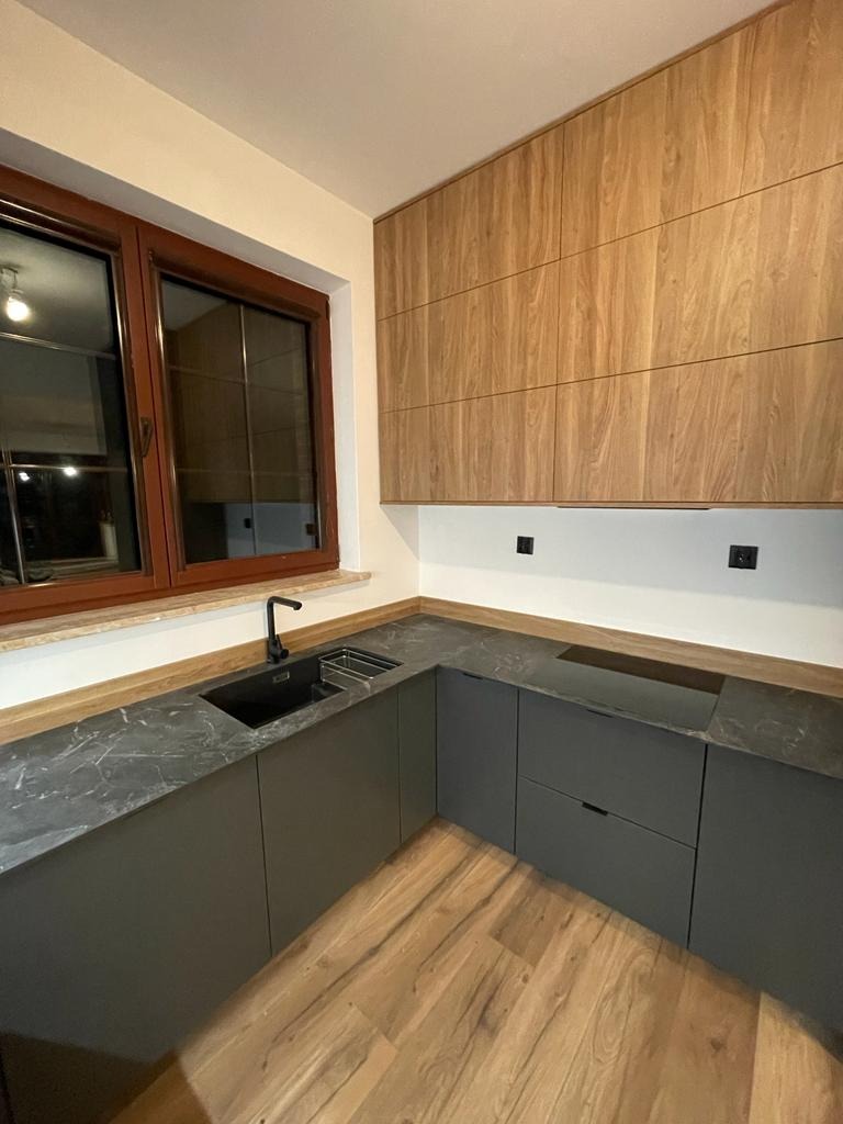 Nowoczesna kuchnia narożna z szarymi szafkami, blatem imitującym marmur i drewnianymi szafkami wiszącymi, czarny zlew i płyta indukcyjna, drewniana podłoga.