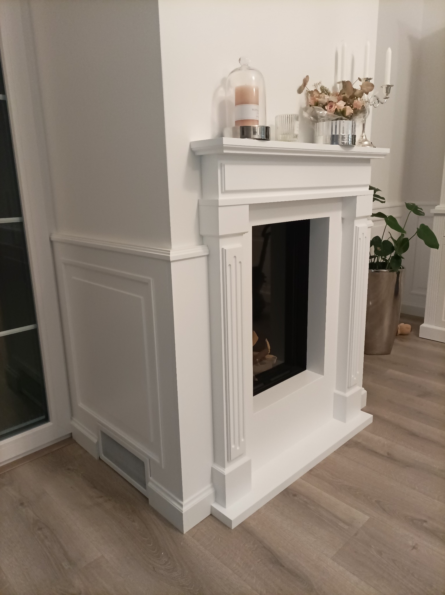 Biały, klasyczny portal kominkowy z dekoracjami na półce, wkomponowany w ścianę z boazerią, widok z boku.