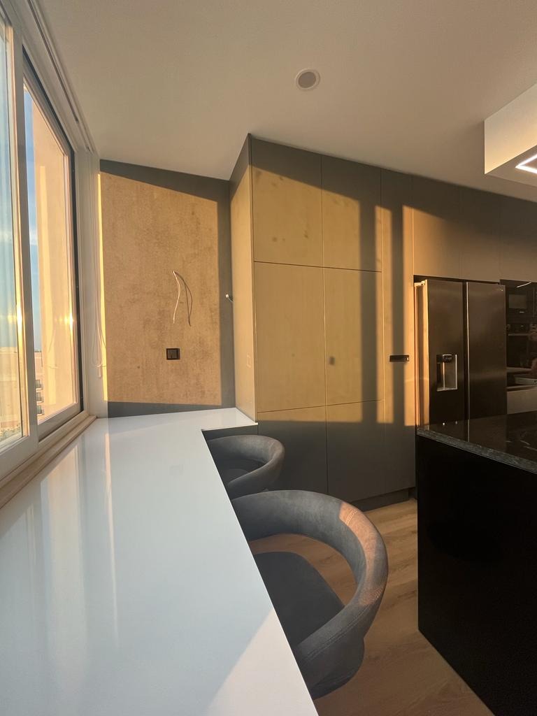 Nowoczesna kuchnia z białym blatem przy oknie, szare szafki i krzesła barowe w odcieniach szarości, oświetlona popołudniowym słońcem.