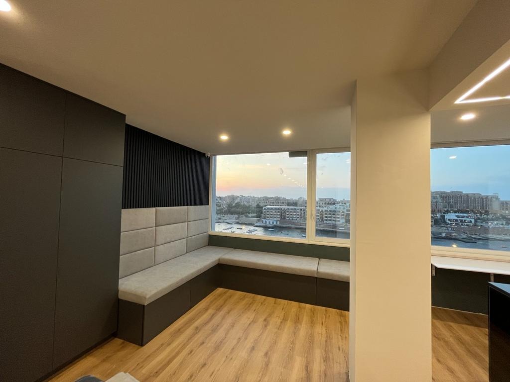 Nowoczesne wnętrze z narożną zabudową siedziska pod oknem, szara tapicerka na ścianie i siedzisku, ciemne drewniane panele pionowe, widok na miasto zza okna.