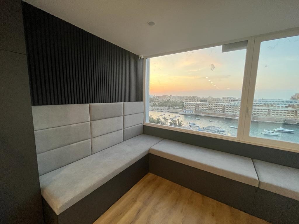 Wnęka okienna z siedziskiem i tapicerowanym panelem ściennym w nowoczesnym stylu, widok na miasto nad morzem o zachodzie słońca.