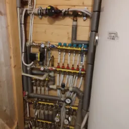 Rozbudowana instalacja hydrauliczna w kotłowni, widoczne rury izolowane szarą pianką, pompy obiegowe Grundfos, rozdzielacze z manometrami i licznymi zaworami, biały zasobnik ciepłej wody, drewniana...