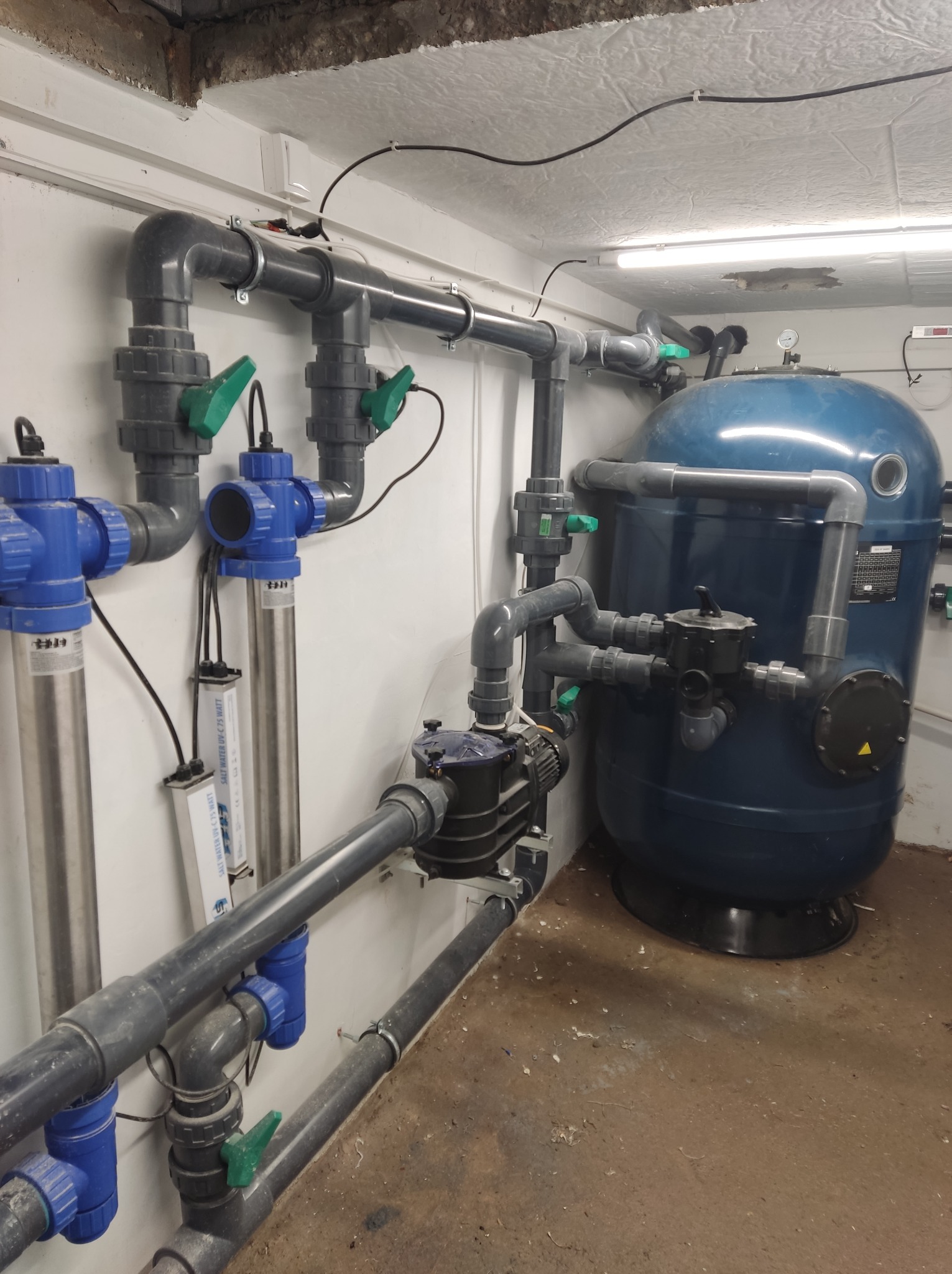 Instalacja filtracyjna wody z niebieskim zbiornikiem, rurami z zaworami i lampami UV-C 75 Watt w pomieszczeniu technicznym.