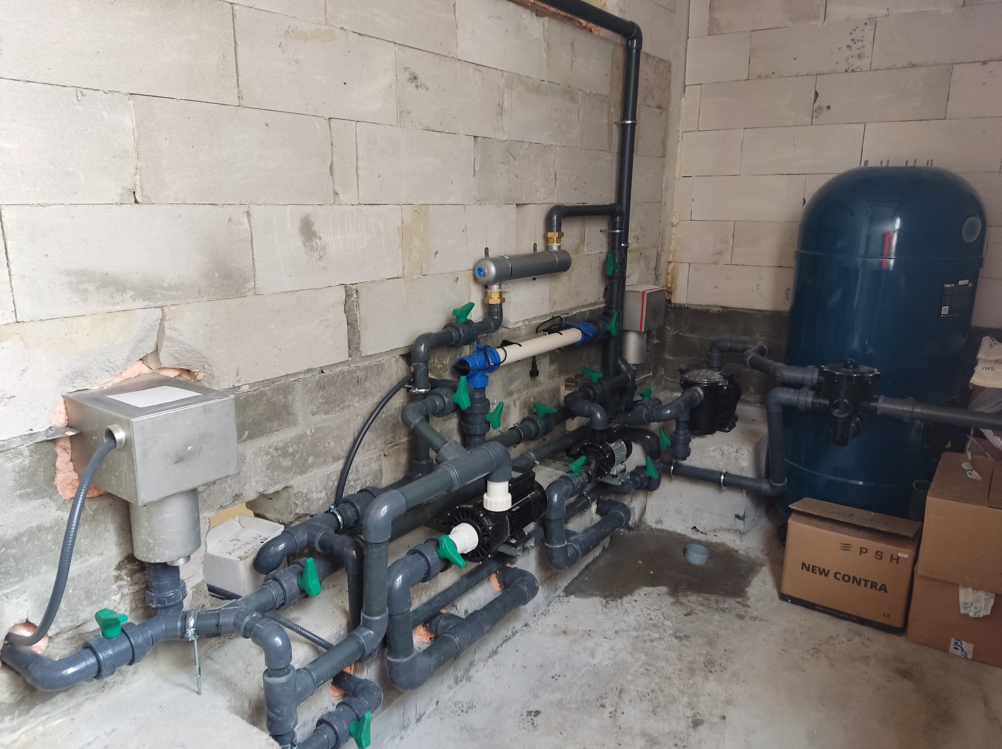 Szare rury instalacji wodnej z zaworami i pompami, zbiornik ciśnieniowy oraz metalowa skrzynka elektryczna na tle ściany z betonu komórkowego. Część rur zabezpieczona izolacją. Widoczne kartony...