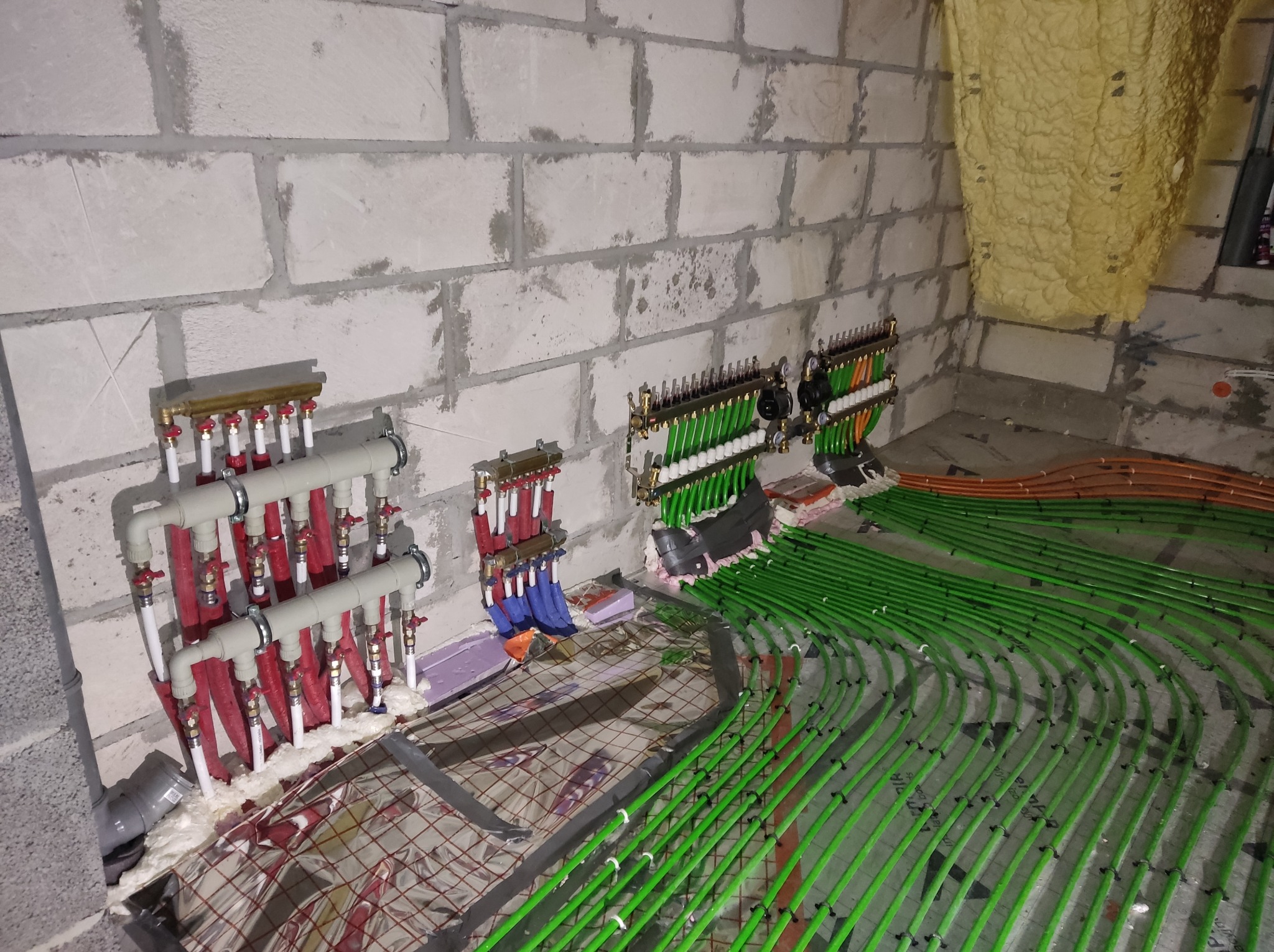 Rozdzielacze instalacji ogrzewania podłogowego z rurami w kolorze zielonym, zamontowane na ścianie z białego bloczka, widoczne fragmenty metalowej siatki i izolacji pod wylewkę.