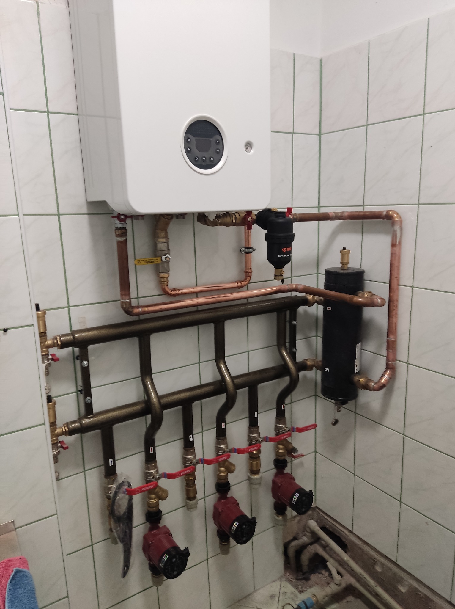 Biały piec grzewczy wiszący na ścianie z białych płytek, z widocznymi miedzianymi rurami, zaworami, filtrami i czerwonymi pompami obiegowymi. Fragment podłogi z elementami instalacji w tle.