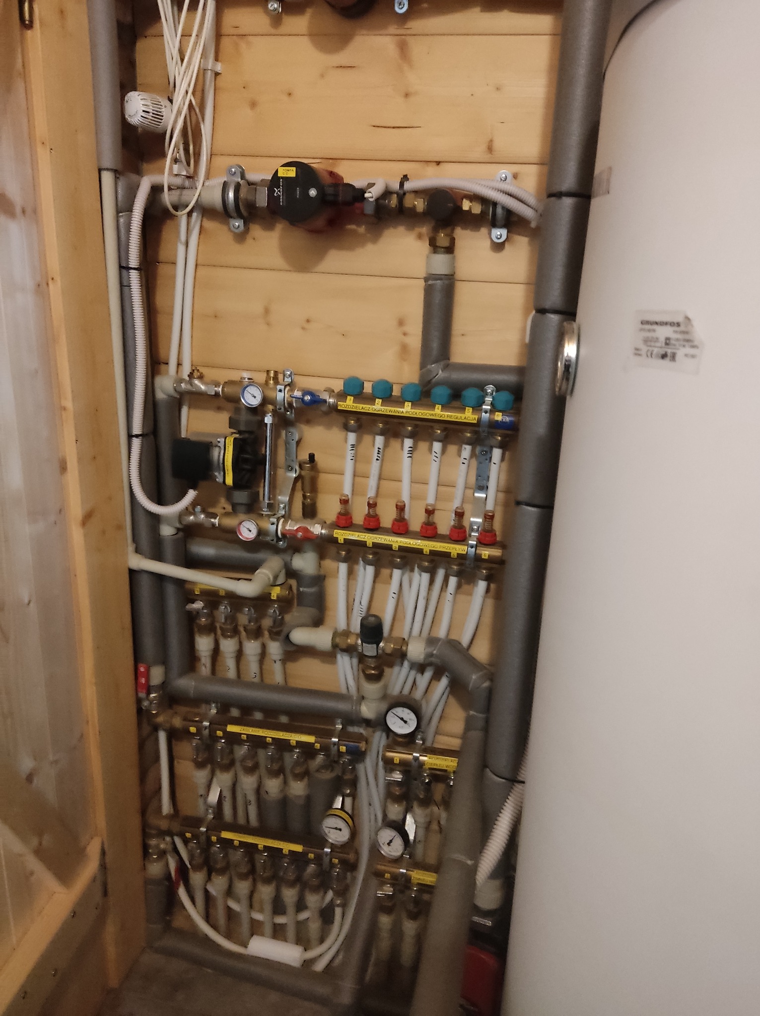 Rozbudowana instalacja hydrauliczna w kotłowni, widoczne rury izolowane szarą pianką, pompy obiegowe Grundfos, rozdzielacze z manometrami i licznymi zaworami, biały zasobnik ciepłej wody, drewniana...