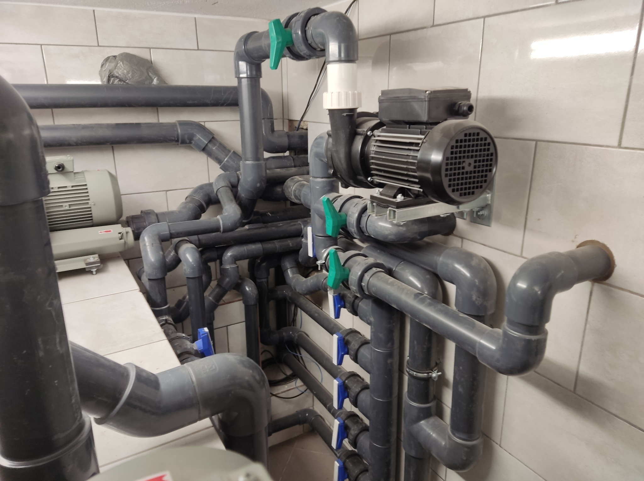 Szare rury PCV, zawory z zielonymi i niebieskimi uchwytami oraz silnik elektryczny na tle białych płytek ceramicznych, instalacja hydrauliczna w kotłowni.