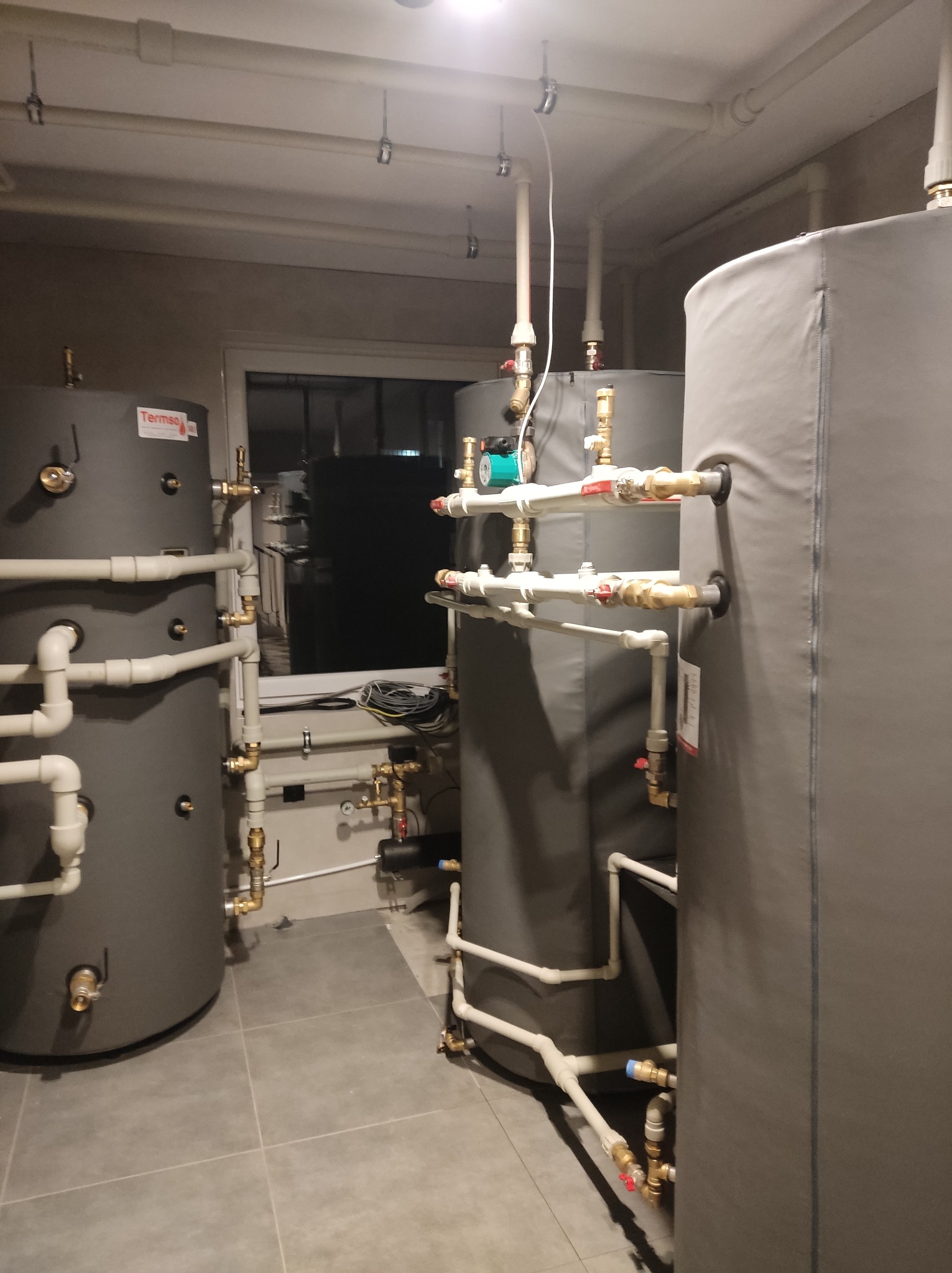 Dwa szare, ocieplone zbiorniki akumulacyjne z licznymi rurami i zaworami w pomieszczeniu z szarą podłogą i oknem w tle. Widoczna pompa obiegowa.