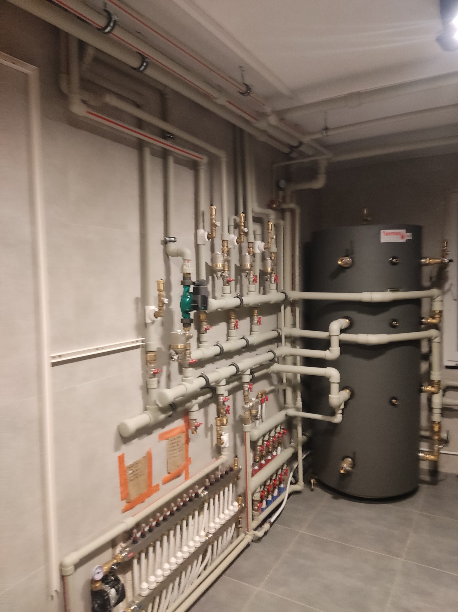 Szara kotłownia z zbiornikiem akumulacyjnym Termoton i rozdzielaczem ogrzewania podłogowego. Instalacja hydrauliczna z licznymi rurami i zaworami.
