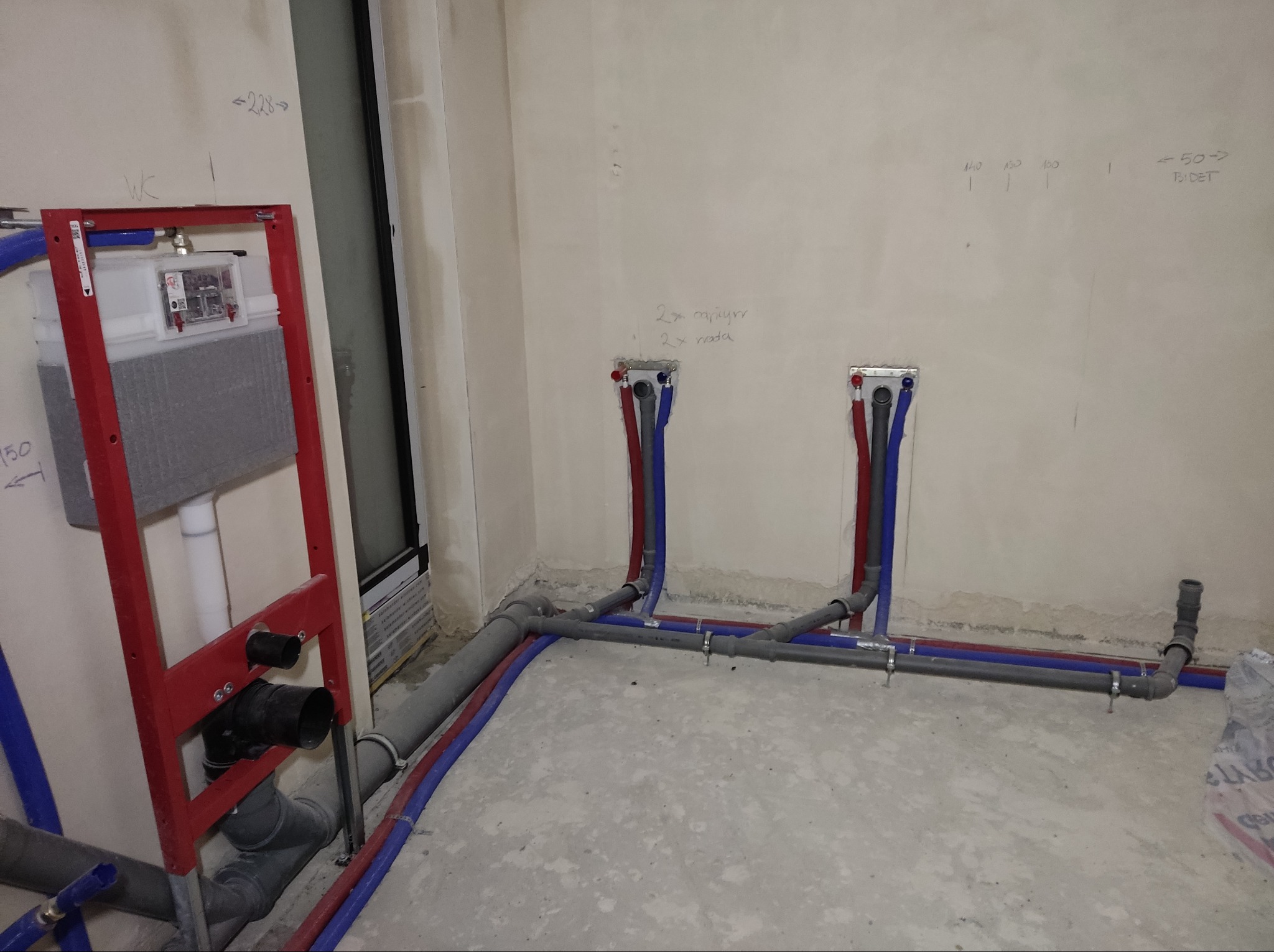 Instalacja podtynkowa WC w stelażu z czerwonym profilem, widoczne rury doprowadzające wodę (czerwone i niebieskie) oraz szare rury kanalizacyjne, surowy stan wykończenia pomieszczenia z widocznymi...
